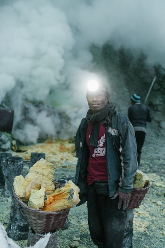 A sulfur miner wearing a headlamp stands near baskets of sulfur amidst volcanic smoke.