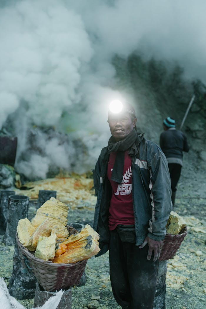 A sulfur miner wearing a headlamp stands near baskets of sulfur amidst volcanic smoke.